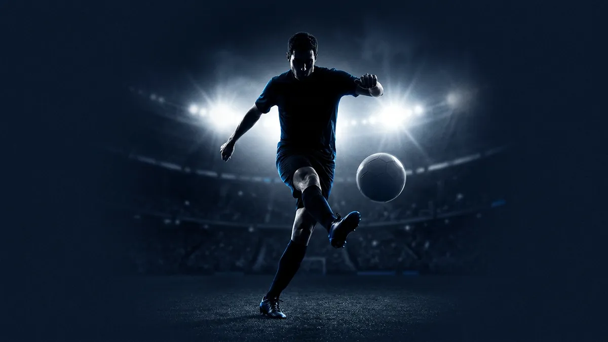 A soccer player in a dark blue kit kicks a ball mid-action on a floodlit stadium pitch at night, silhouetted against blazing white stadium lights with a blurred crowd in the background.