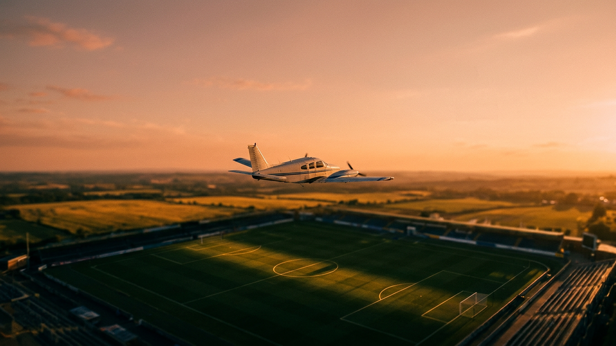 A small propeller plane silhouetted against a vast open sky, casting a long shadow over an abstracted football pitch below