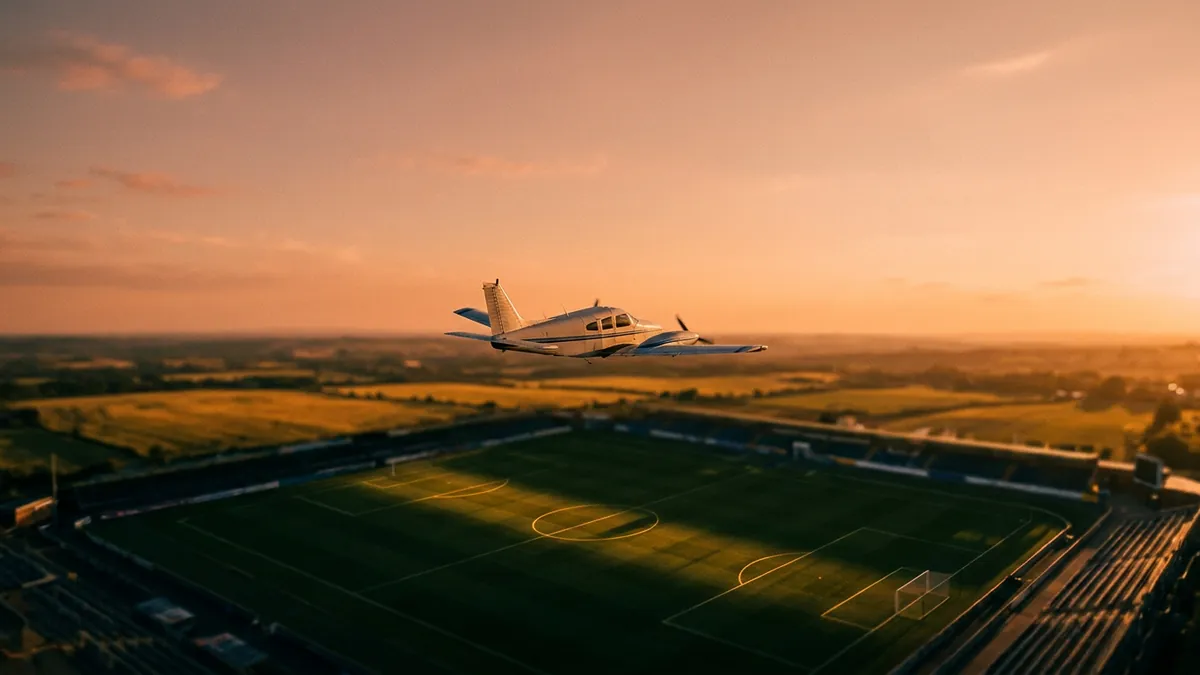 A small white twin-engine propeller plane flies low over a football stadium at golden-hour sunset, with green pitch markings visible below and rural fields stretching to the warm orange horizon.