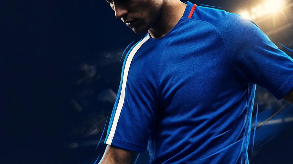 A male athlete in a royal blue short-sleeve football jersey with a white stripe and red trim leans forward under bright stadium floodlights against a dark blue background.