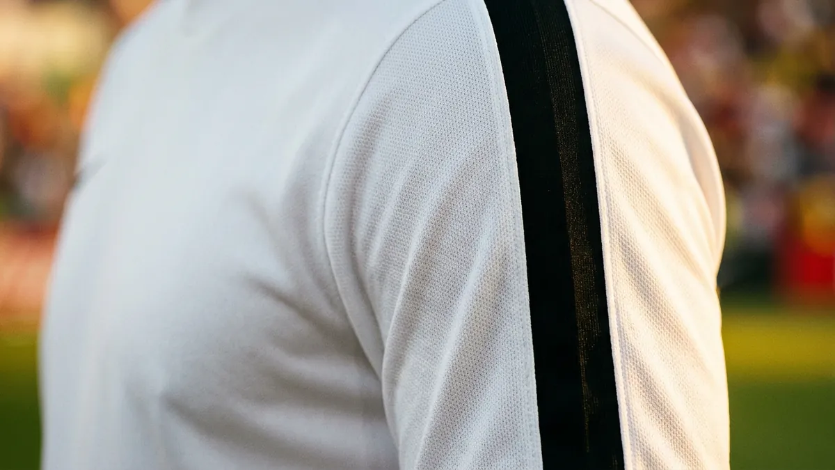 Close-up of a person's upper back and shoulder wearing a white athletic jersey with a vertical black stripe running down the outer sleeve, set against a blurred green pitch and warm-toned crowd.