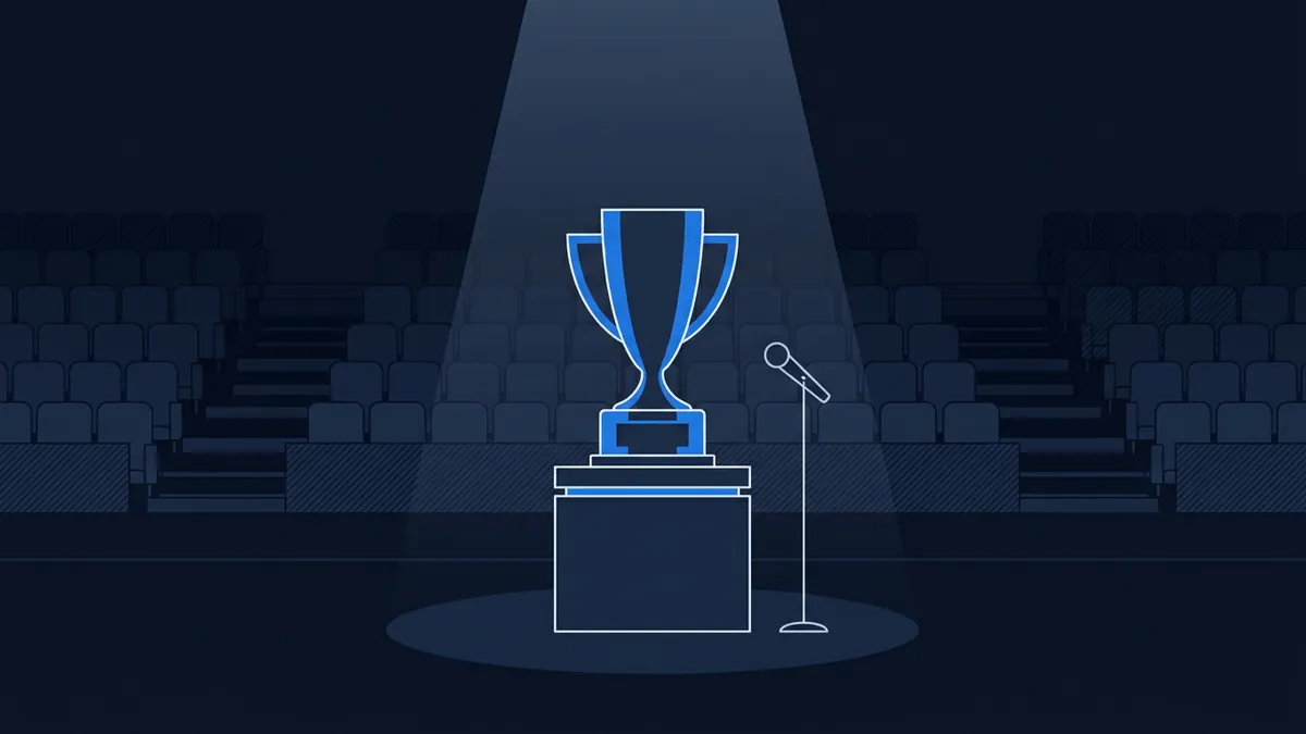 A blue trophy cup sits on a pedestal under a spotlight on an empty stage, with a microphone stand beside it and rows of dark empty seats in the background.