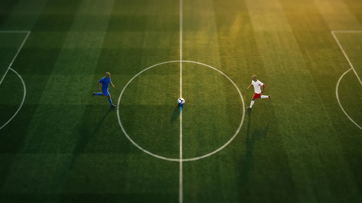 Aerial view of a green football pitch at the centre circle, where one player in blue kit and one in red and white kit run toward a soccer ball, bathed in warm golden light.
