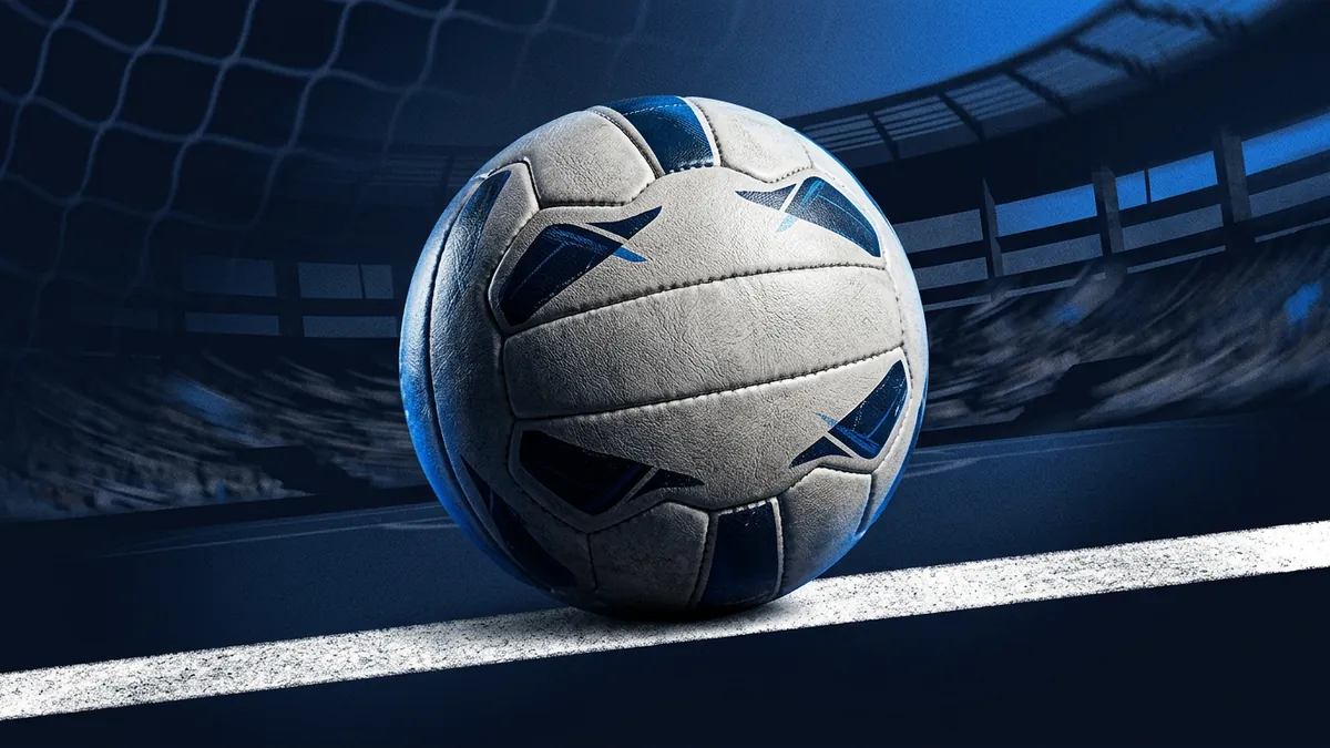 A white and navy leather football sits on a painted white goal-line, with a blurred stadium structure and goal net visible in a deep blue background.