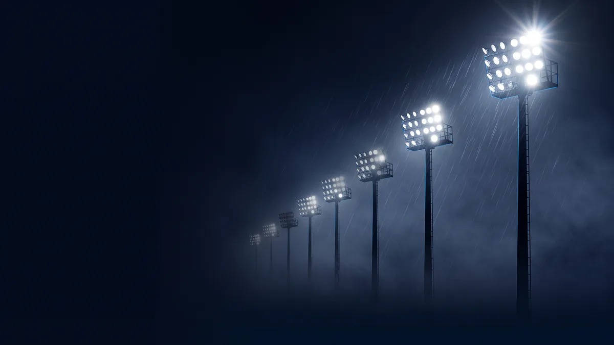 A row of eight stadium floodlight banks on tall masts at night, seen from pitch level and receding diagonally into blue mist, the nearest blazing white with a lens starburst and the farthest almost dark, rain streaking across the light; the left third of the frame is empty near-black navy.