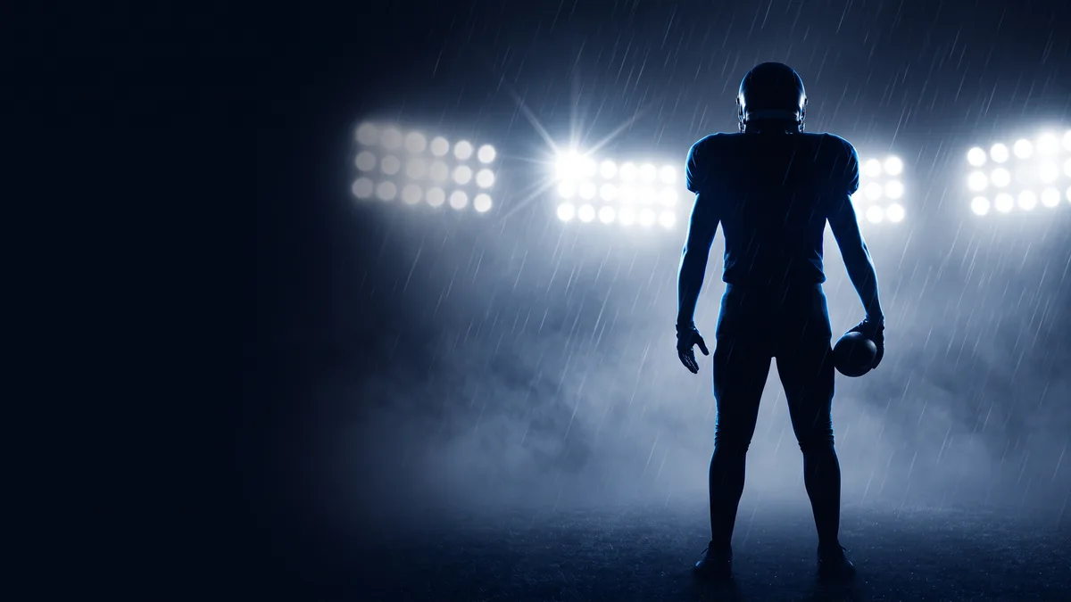 A lone American football player in full silhouette seen from behind, standing in rain with a ball at his hip, facing a bank of white stadium floodlights that rim his helmet and shoulders in cold blue light, mist filling the ground below; the left third of the frame is empty near-black navy.