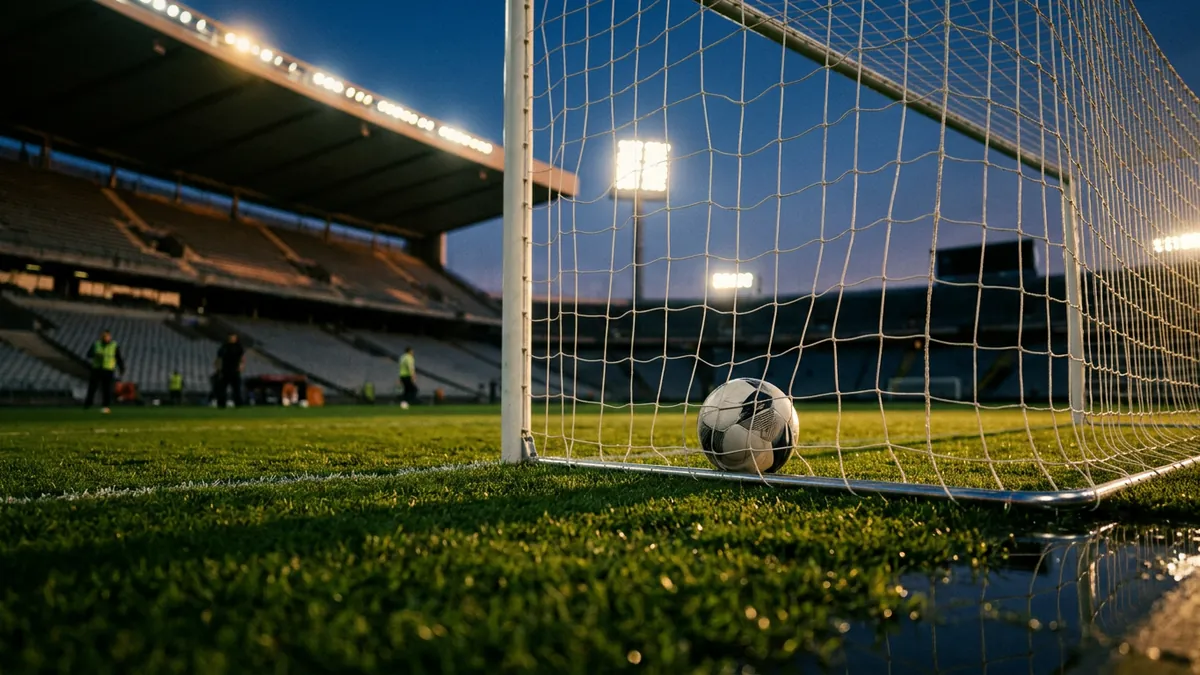 A football sits inside a goal net at ground level in an empty stadium at dusk, with floodlights glowing blue and three figures visible on the pitch in the background.